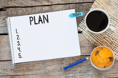 On a rustic slatted wood table, a small blue pencil, a blueberry muffin, a cup of coffee on a woven mat, and a notebook open to a white page titled PLAN in black, with five numbered but blank rows representing bankruptcy planning for non-exempt assets in MN.