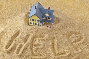 A small, yellow, 2-story model house with a blue-grey roof falling into beach sand on which the word "HELP" have been written with a finger, posing the question, Can you file bankruptcy and keep your home in Minnesota?