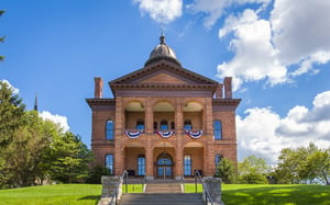 A picture of a Minnesota court house surrounded by green grass and trees, against a blue sky with white clouds, posing the question, Do I have to go to court when I file bankruptcy in Minnesota during the COVID-19??