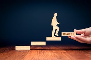 A hand holds a small wooden rectangle cutout with the word QUALIFIED on it, which is the fourth floating step in the series. A small wooden cutout of a man is walking up the floating steps, towards the QUALIFIED step, raising the question, How do I qualify to file a chapter 7 bankruptcy?