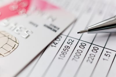 A white credit card and silver pen tip sit on top of a bank statement listing deposit and withdrawal amounts, posing the question, How much is too much money in a bank account during bankruptcy in MN?
