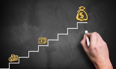 A hand draws on the far right side of a chalkboard, creating a simple staircase from left to right in white, and three images in yellow of a stack of coins, stack of paper currency, and a bag of money with a $ sign. The coins are on the first step, the paper currency on the fourth, and money bag on the eighth. The scene represents the question, If my income increases after the pandemic, will it affect my bankruptcy?