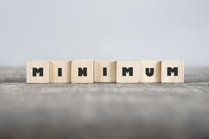 Seven light wood, scrabble-type tiles, each with a thick black letter, spelling out "MINIMUM". The tiles sit on a grey wood-like table, posing the question, Is there a minimum debt requirement to file bankruptcy in Minnesota?