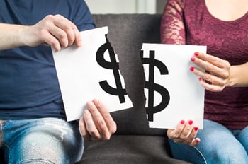 A man and a woman sit next to each other on a grey couch, each holding one-half of a torn white piece of paper with a black dollar sign on it, representing limiting the impact of the bankruptcy on the non-filing spouse.