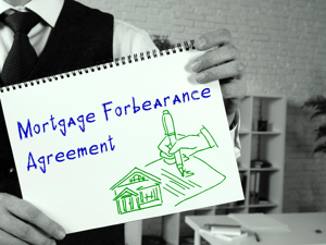 A man in a suit sans jacket holds a sketchbook up with a picture in green of a hand signing an agreement next to a small bank, and in blue the words "Mortgage Forbearance Agreement".