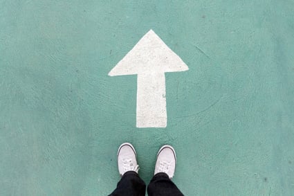 Legs from the ankle down in black pants and white tennis shoes, at the bottom center of the image are standing in front of a large white arrow painted on mint green concrete, representing taking that first step towards filing MN bankruptcy.