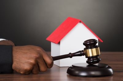 A hand strikes a block with a dark-wood gavel in front of a small white toy house with a red roof, all placed on a wood table to represent understanding Minnesota law to protect your property.