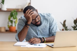 A black man wearing a blue shirt is holding his head in his right hand in exasperation while sitting at a desk with a laptop and financial papers for filing bankruptcy asks the question, What are the risks of filing bankruptcy without a lawyer?