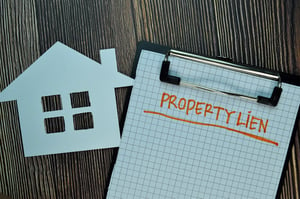 A white paper cut-out of a house and a piece of grid paper on a  clipboard on a wooden table. The paper has "Property Lein" written on it in orange marker with an underline, posing the question, Why do I have a tax lien on my home in Minnesota?