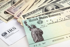 Dollar bills in increments of $50 and $100 are strewn on a white table next to a United States Treasury check and piece of paper that says "IRS", posing the question, Will I Lose My Tax Refund If I File Chapter 7 in Minnesota?