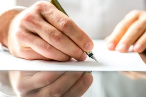 A hand holding a pen as it writes onto a Bankruptcy Means Test on a white piece of paper on a reflective glass desk.