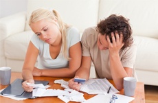 A young couple is asking, Should I stop paying my bills before bankruptcy in Minnesota?, as they are sitting in front of a white sofa with bills in front of them on the coffee table.