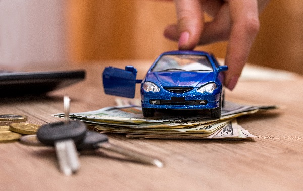 A hand touches a blue toy car with an open door as it sits atop a stack of money and next to a set of car keys on a light wood table, posing the question, Can filing bankruptcy in MN reduce my auto loan payments?