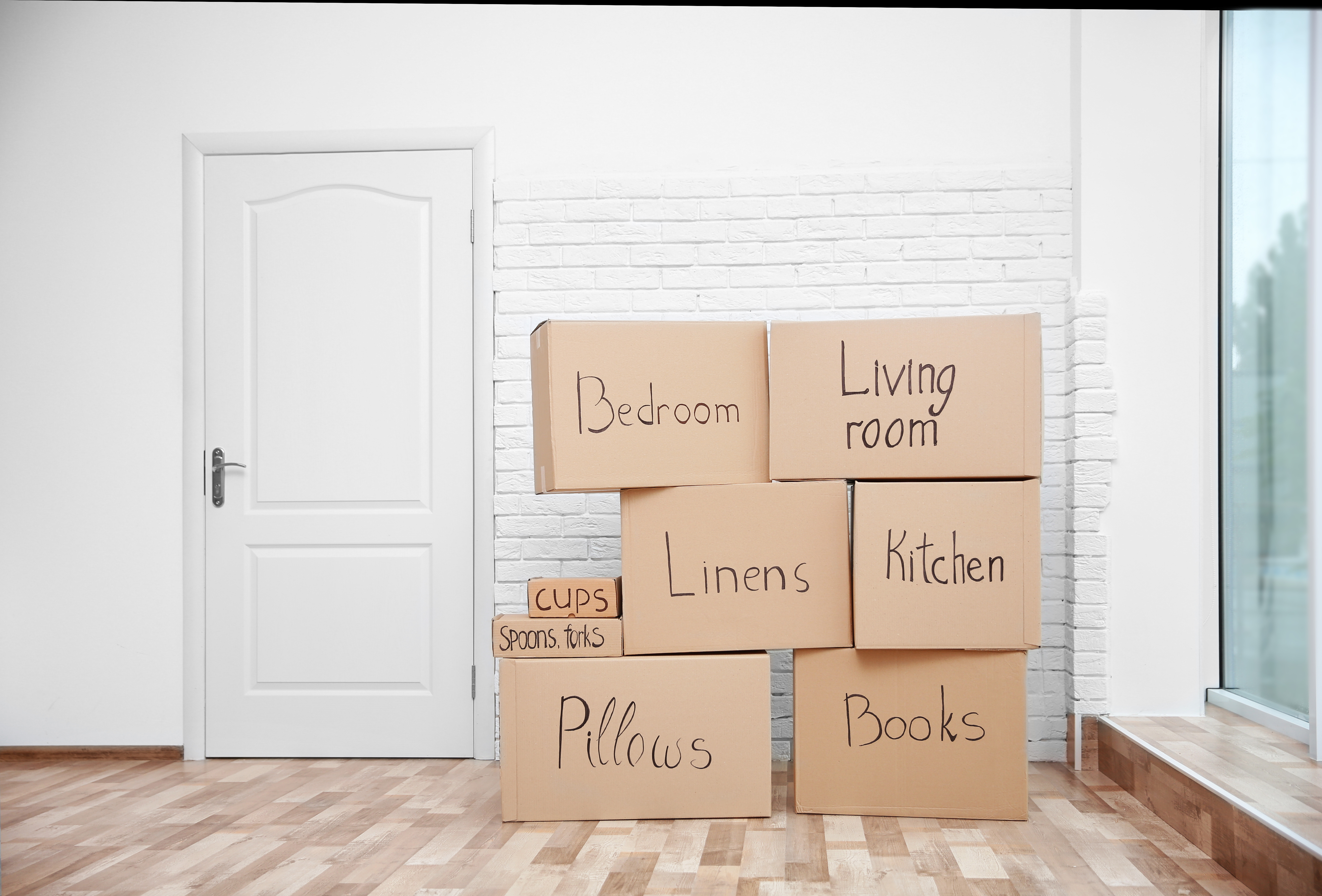 Moving boxes with contents marked in black marker sit faux light wood flooring and in front of a white wall with some brick, with a white door, posing the question, Can I file bankruptcy in Maple Grove MN if I only recently moved here?