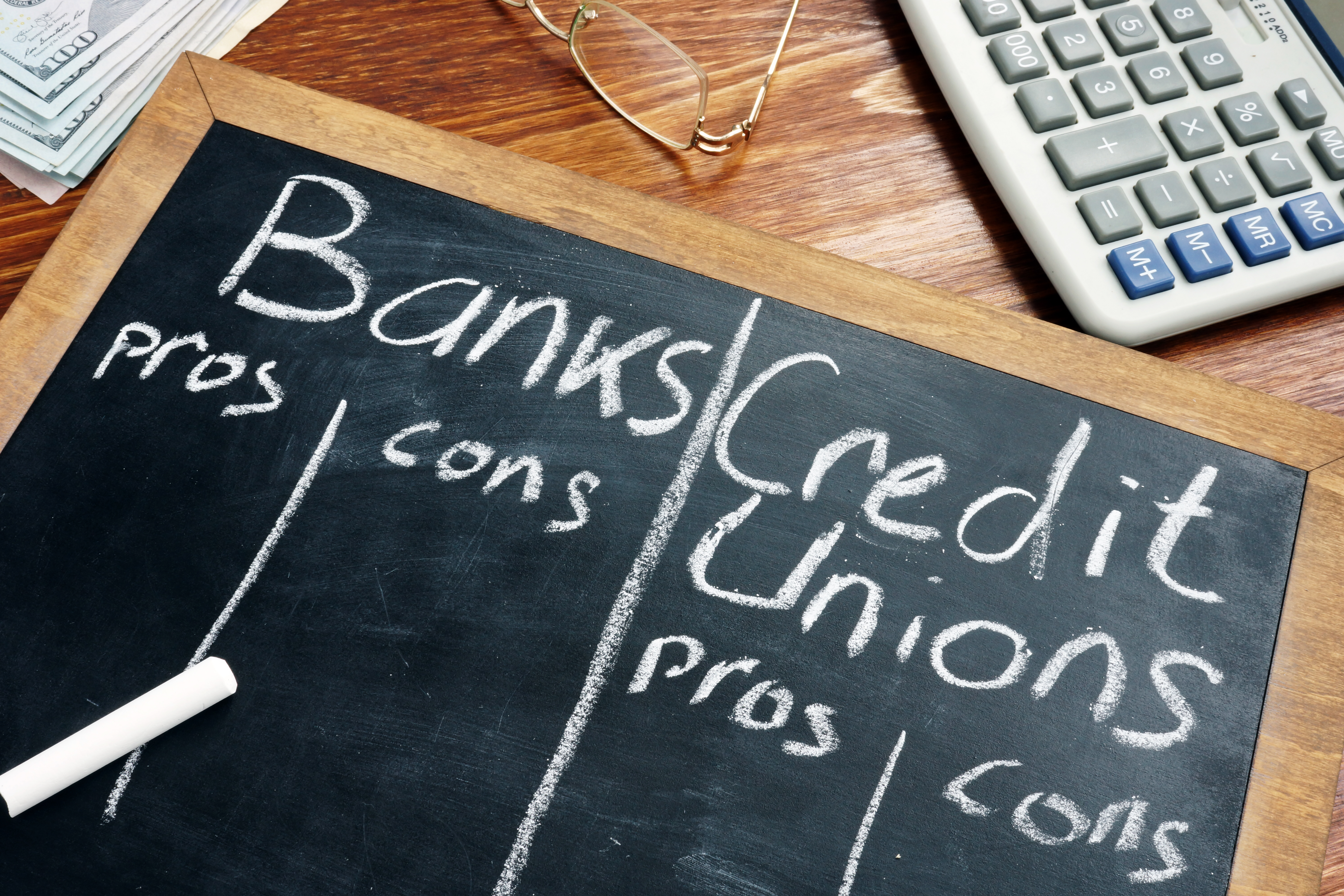A wood table with a calculator, reading glasses, a stack of white papers surrounding a wood framed chalk board that takes up most of the image, with two columns, titled Banks and Credit Unions and drawn in white chalk. The two main columns each have Pros and Cons columns that are empty, representing what you need to know about credit unions and filing bankruptcy.