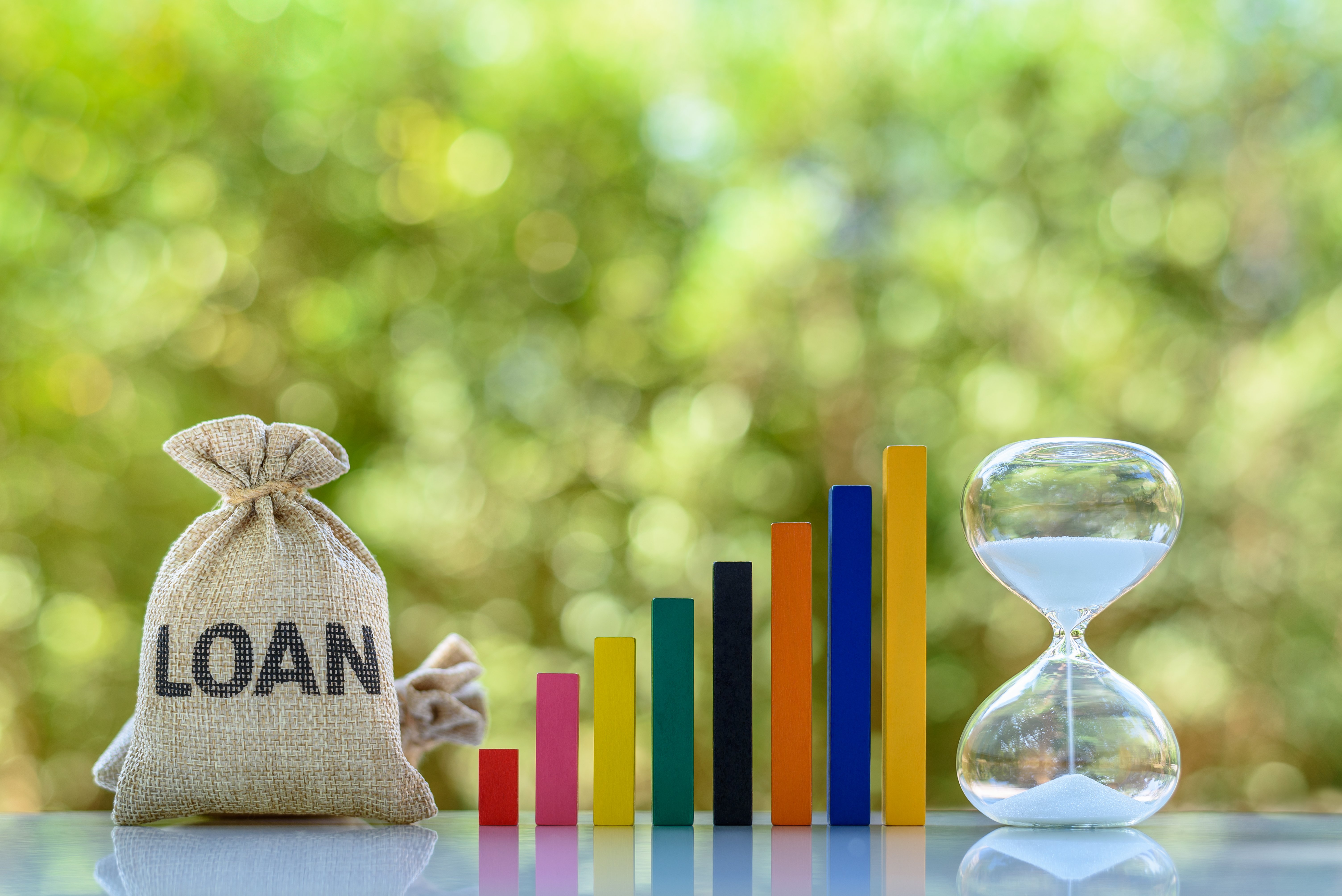 Placed on a gray table with a green foliage background are from left to right: two burlap sacks with LOAN written on them in black, eight multi-colored wooden rectangular pieces graduating in height to form a bar graph, and an hourglass with flowing white sand. These objects symbolize the repayment of loans before filing a bankruptcy in Minnesota.