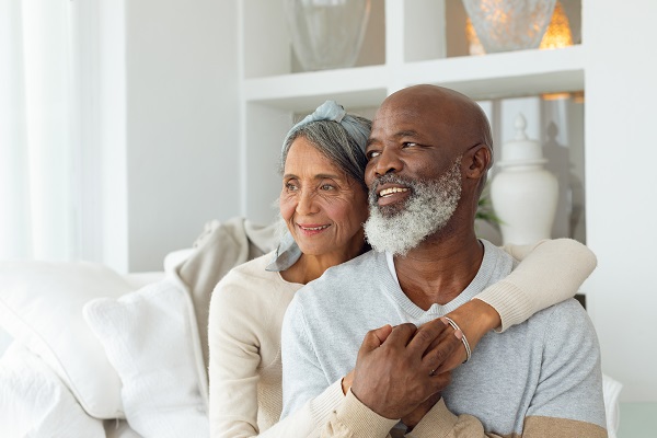 An elderly couple, sitting in their home, the women sitting slightly behind the smiling man with her left arm around his left shoulder as his right hand hold her left. The scene poses the question, Should I use retirement funds to pay my debt in MN?