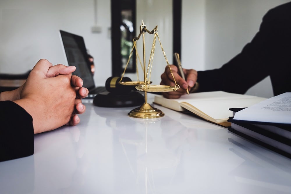 Closeup of a white desk with clasped hands of a debtor on one side, a miniature scales of justice, a laptop, a creditor's hand with pen and notebooks on the other, posing the question, What Do I Say in My Minnesota Bankruptcy 341 Meeting?