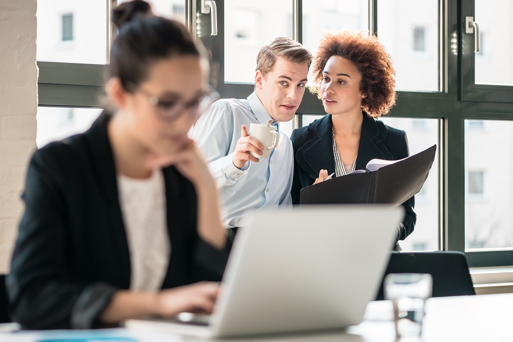 A man and woman gossiping in the background about a woman working on a laptop in the foreground, exemplifying what your co-debtor needs to know when you file bankruptcy in MN