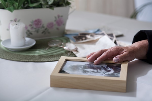 A white candle, green placemat, reading glasses, tabletop houseplant in a white pot, and photos in the background on a white tabletop. In the foreground, the hand of an elderly person gently touches a black and white photo of their deceased male spouse, framed in light wood. The question posed is, Will my surviving spouse have to pay my debts if I die in Minnesota?